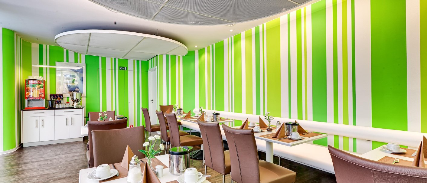Moderner Frühstücksraum mit grünen Streifenwänden, gedeckten Tischen und braunen Stühlen. Ein Getränkeautomat steht in der Ecke., © attimo Hotel Stuttgart Moderner Frühstücksraum mit grünen Streifenwänden, gedeckten Tischen und braunen Stühlen. Ein Getränkeautomat steht in der Ecke., © attimo Hotel Stuttgart