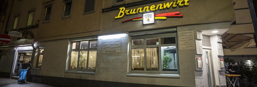 Das Restaurant Brunnenwirt bei Nacht, beleuchtet mit gelbem Schriftzug. Ein Mann steht an einem Tisch vor dem Eingang., © SMG, Sarah Schmid Das Restaurant Brunnenwirt bei Nacht, beleuchtet mit gelbem Schriftzug. Ein Mann steht an einem Tisch vor dem Eingang., © SMG, Sarah Schmid