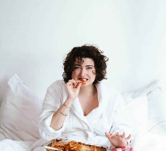 Eine Person im wei&szlig;en Bademantel sitzt im Bett und isst Lasagne aus einer Auflaufform. Sie h&auml;lt ein St&uuml;ck Lasagne in der Hand., &copy; Rosenau Kultur e.V.