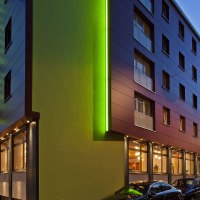 Modern hotel exterior view at dusk, with green lighting and parked cars. The lettering 'Hotel Wartburg' is visible., © VCH Hotel Wartburg Modern hotel exterior view at dusk, with green lighting and parked cars. The lettering 'Hotel Wartburg' is visible., © VCH Hotel Wartburg