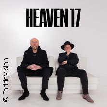 Zwei Männer in dunkler Kleidung sitzen auf einem weißen Sofa. Im Hintergrund steht 'Heaven 17'., © links im Bild