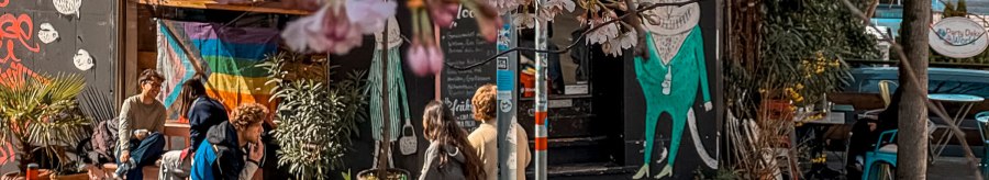 A cozy street café with a yellow sunshade, rainbow flag and blooming cherry blossoms. People sit relaxed in front of it., © Café Moody, Stuttgart A cozy street café with a yellow sunshade, rainbow flag and blooming cherry blossoms. People sit relaxed in front of it., © Café Moody, Stuttgart
