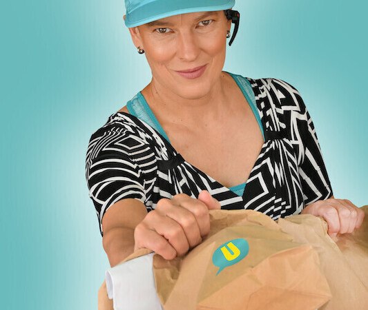 Woman with blue visor and headset hands a paper bag with logo in front of a turquoise background., &copy; Renitenztheater Stuttgart e.V.