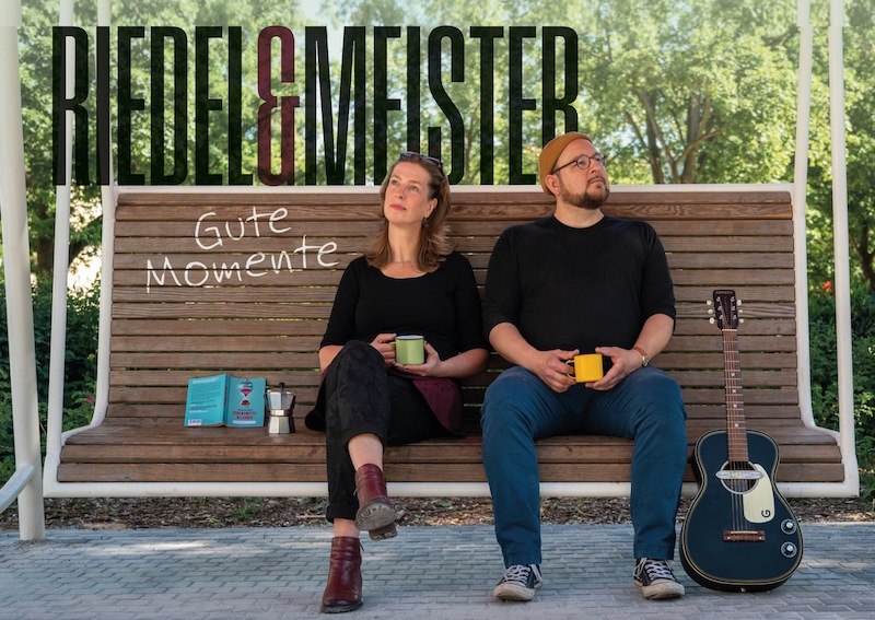 Two people sit on a bench with coffee cups, a guitar stands next to them. In the background is the lettering 'Riedel & Meister' and 'Gute Momente'., &copy; Rosenau Kultur e.V.