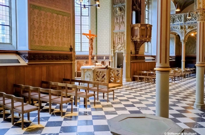 Die Schlosskirche Stuttgart zeigt eine elegante Innenansicht mit Holzbänken, einem Kruzifix und einer kunstvoll geschnitzten Kanzel auf einem schwarz-weißen Boden., © Stiftsmusik Stuttgart Die Schlosskirche Stuttgart zeigt eine elegante Innenansicht mit Holzbänken, einem Kruzifix und einer kunstvoll geschnitzten Kanzel auf einem schwarz-weißen Boden., © Stiftsmusik Stuttgart