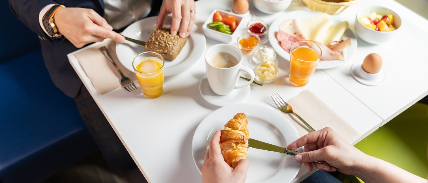 Zwei Personen frühstücken mit Croissants, Brötchen, Aufschnitt, Käse, Eiern, Joghurt und Orangensaft an einem Tisch., © DQuadrat Living GmbH Zwei Personen frühstücken mit Croissants, Brötchen, Aufschnitt, Käse, Eiern, Joghurt und Orangensaft an einem Tisch., © DQuadrat Living GmbH