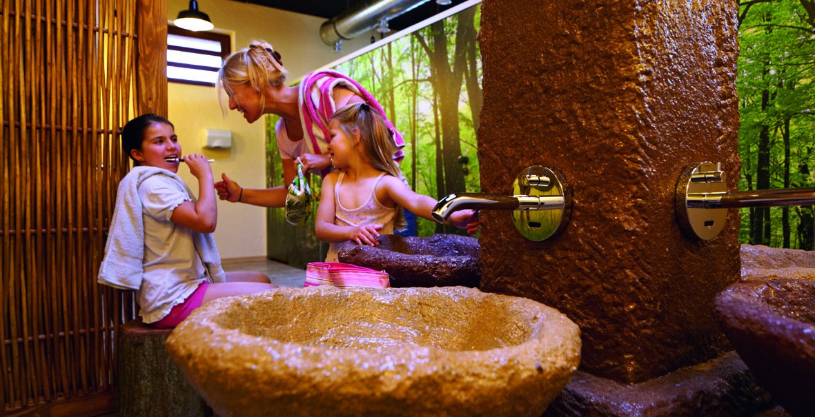 Drei Personen in einem Badehaus mit Naturdekor. Eine Frau und zwei Kinder interagieren fröhlich neben einem steinernen Waschbecken., © Tripsdrill Natur-Resort GmbH Drei Personen in einem Badehaus mit Naturdekor. Eine Frau und zwei Kinder interagieren fröhlich neben einem steinernen Waschbecken., © Tripsdrill Natur-Resort GmbH