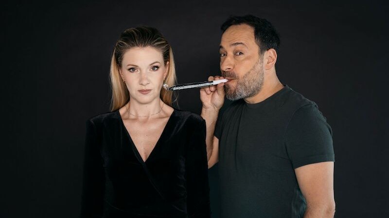 A woman in a black dress looks serious while a man next to her blows a party horn. The background is black., &copy; Theaterhaus Stuttgart e.V.