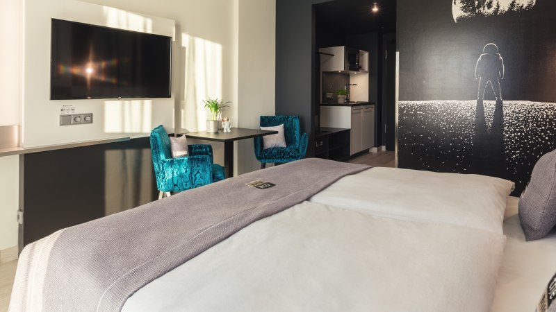 Modernes Zimmer mit großem Bett, blauen Sesseln, Astronauten-Wandbild, Flachbildfernseher und kleiner Küche., © Novum Management GmbH Modernes Zimmer mit großem Bett, blauen Sesseln, Astronauten-Wandbild, Flachbildfernseher und kleiner Küche., © Novum Management GmbH