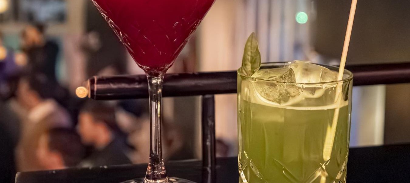 Zwei Cocktails auf einer Bar: ein roter im Martiniglas und ein grüner im Tumblerglas mit Strohhalm, im Hintergrund unscharfe Bar-Szene., © SMG, Sarah Schmid