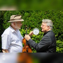 Zwei Männer stehen sich gegenüber, einer mit Geige, der andere mit einer Uhr. Sie diskutieren im Freien vor grünen Büschen., © links im Bild Zwei Männer stehen sich gegenüber, einer mit Geige, der andere mit einer Uhr. Sie diskutieren im Freien vor grünen Büschen., © links im Bild