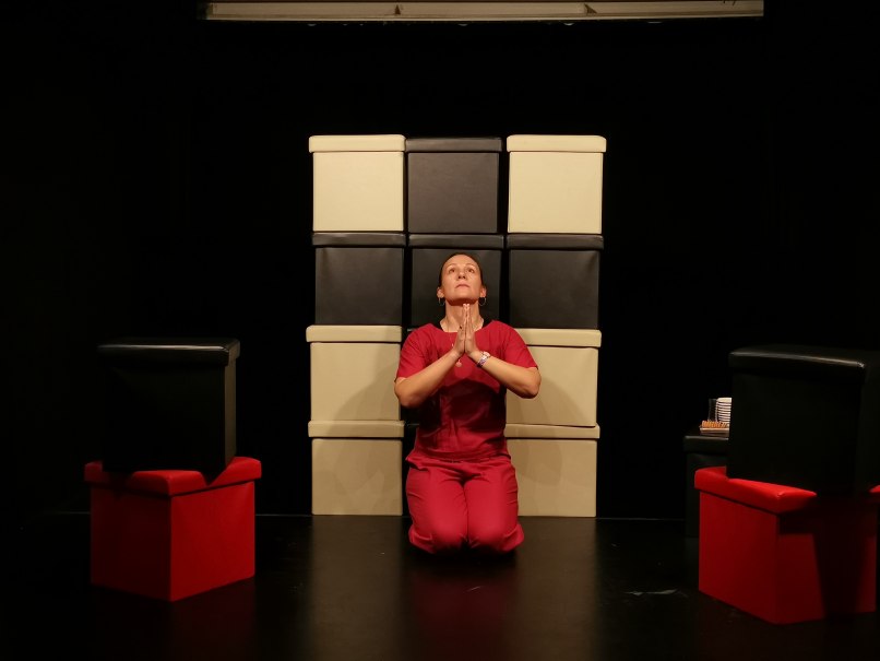 Person in roter Kleidung kniet vor gestapelten Boxen, Hände betend gefaltet. Dunkler Hintergrund, rote und schwarze Boxen., © Maja Kles