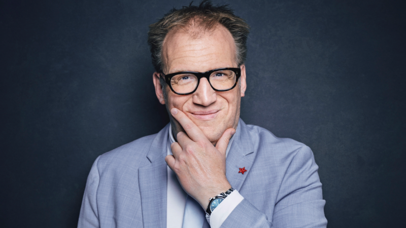 Ein Mann in einem hellen Anzug mit Brille lächelt und berührt nachdenklich sein Kinn vor einem dunklen Hintergrund., © Olli Haas Ein Mann in einem hellen Anzug mit Brille lächelt und berührt nachdenklich sein Kinn vor einem dunklen Hintergrund., © Olli Haas