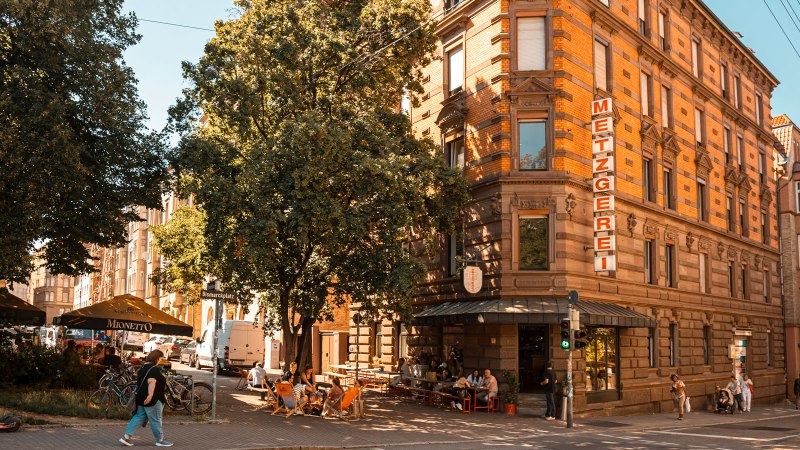 Ein belebter Platz mit einer Metzgerei, Caf&eacute;g&auml;sten unter Sonnenschirmen und einem gr&uuml;nen Ampellicht. Menschen spazieren und entspannen sich., &copy; SMG, Sarah Schmid