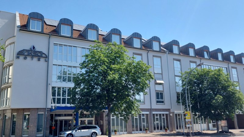 Moderne Hotelfassade mit großen Fenstern und Dachgauben, ein Baum und ein Auto davor. Klare, sonnige Wetterbedingungen., © Erikson Hotel