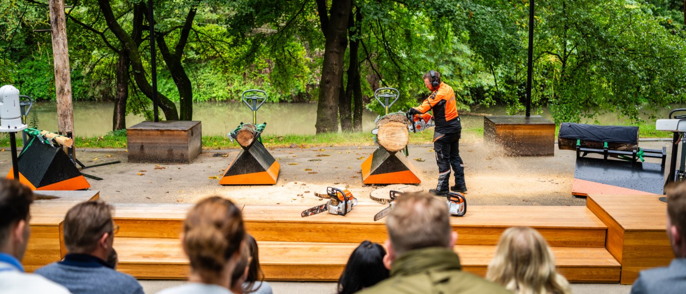 An outdoor demonstration area where a person wearing protective gear is demonstrating how to use a chainsaw on a log. Spectators are watching the demonstration., © STIHL Markenwelt An outdoor demonstration area where a person wearing protective gear is demonstrating how to use a chainsaw on a log. Spectators are watching the demonstration., © STIHL Markenwelt