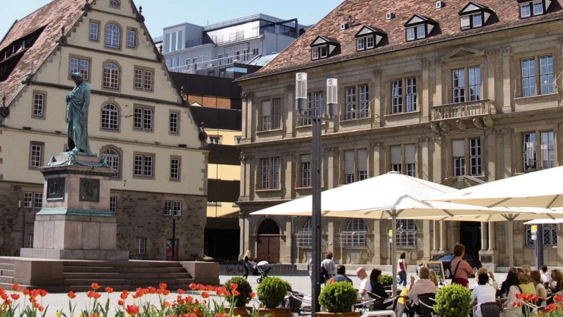 Der Schillerplatz in Stuttgart mit einer Statue im Vordergrund, umgeben von historischen Geb&auml;uden und Menschen, die unter Sonnenschirmen sitzen., &copy; Stuttgart-Marketing GmbH