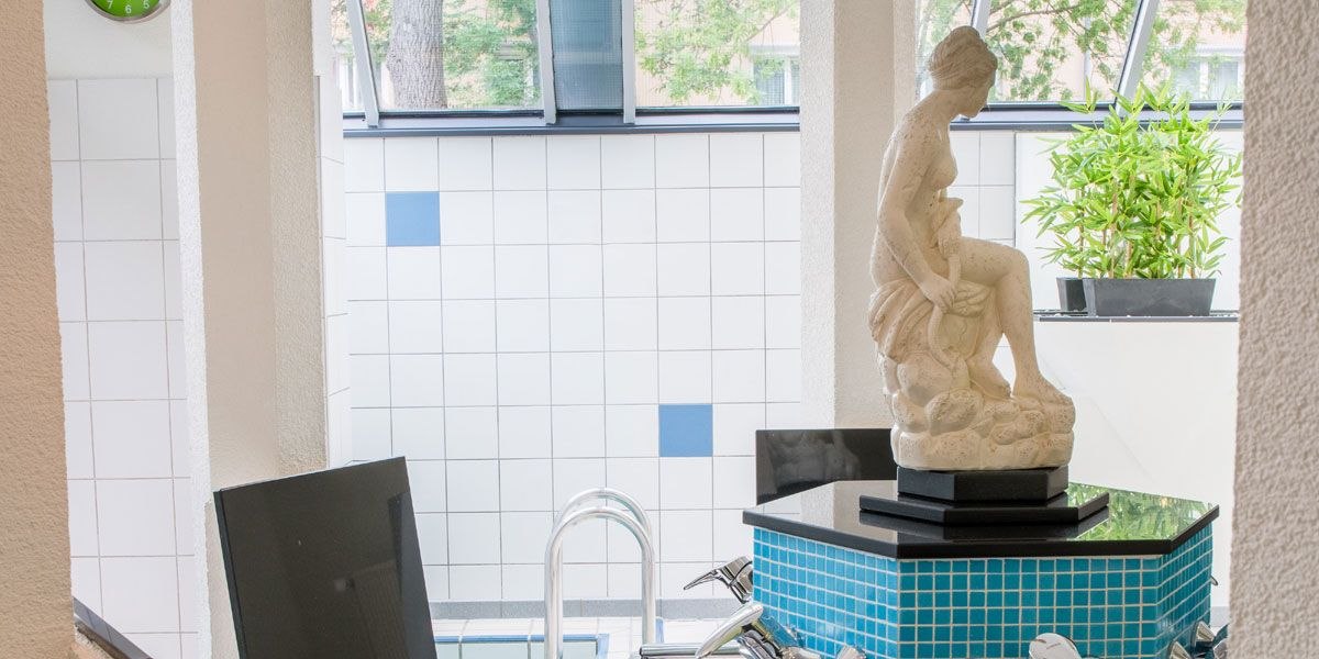 Ein Spa-Bereich mit einer Statue auf einem blauen Mosaiktisch, umgeben von weißen Fliesen. Im Hintergrund sind Pflanzen und ein Fenster zu sehen., © Best Western Plus Hotel am Schlossberg