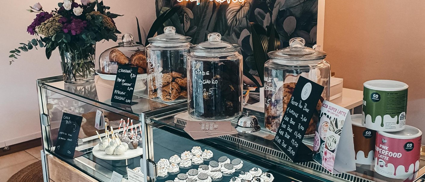 Ein Café mit einer Glasvitrine voller Cupcakes und Kekse. Ein Neon-Schild mit 'Cupcake' leuchtet im Hintergrund. Blumen schmücken den Tresen., © Cupcakes & Bagels, Stuttgart Ein Café mit einer Glasvitrine voller Cupcakes und Kekse. Ein Neon-Schild mit 'Cupcake' leuchtet im Hintergrund. Blumen schmücken den Tresen., © Cupcakes & Bagels, Stuttgart