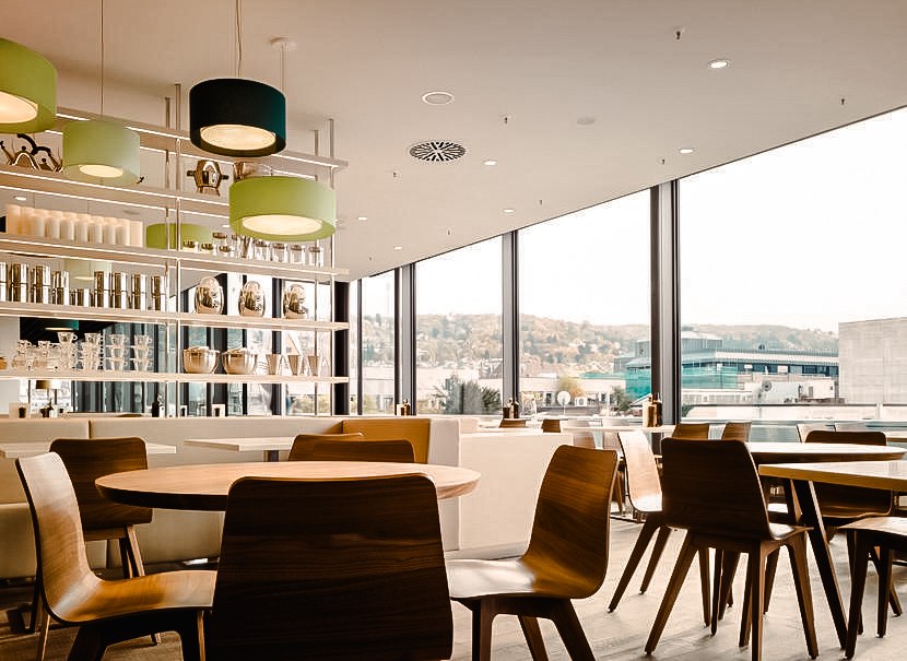 Modernes Café mit Holzmöbeln, großen Fenstern und Blick auf Stuttgart. Helle Lampen und Regale mit Geschirr dekorieren den Raum., © Breuninger
