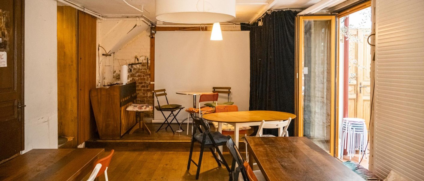 Gemütliches Café mit Holztischen, Stühlen und warmem Licht. Eine Tür führt nach draußen, im Hintergrund sind gestapelte Hocker zu sehen., © SMG, Sarah Schmid