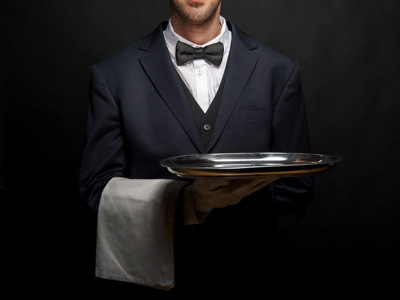 A waiter in a suit and bow tie presents a silver tray against a dark background. He wears a white scarf over his arm., &copy; engesser marketing GmbH