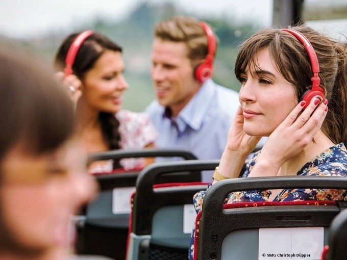 Menschen sitzen in einem Tourbus und tragen rote Kopfhörer. Sie genießen die Audiotour während der Fahrt durch die Stadt., © SMG, Christoph Düpper Menschen sitzen in einem Tourbus und tragen rote Kopfhörer. Sie genießen die Audiotour während der Fahrt durch die Stadt., © SMG, Christoph Düpper