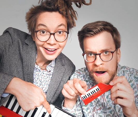 Zwei Personen mit Brillen posieren fröhlich mit einem roten Keyboard und einer Miniaturtastatur. Die Szene wirkt humorvoll und kreativ., © Rosenau Kultur e.V. Zwei Personen mit Brillen posieren fröhlich mit einem roten Keyboard und einer Miniaturtastatur. Die Szene wirkt humorvoll und kreativ., © Rosenau Kultur e.V.