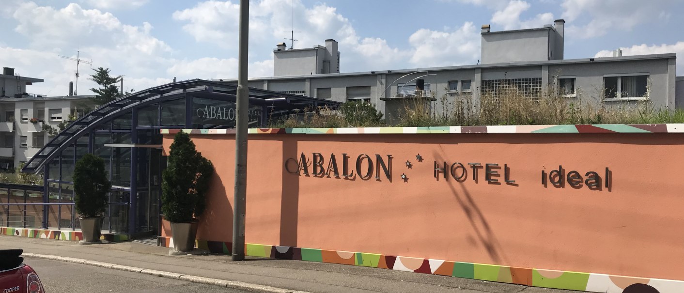 Außenansicht eines Hotels mit orangefarbener Wand, Glasdach und Pflanzen. Schriftzug 'ABALON HOTEL ideal' sichtbar. Straße und Auto im Vordergrund., © Abalon Hotel Ideal Außenansicht eines Hotels mit orangefarbener Wand, Glasdach und Pflanzen. Schriftzug 'ABALON HOTEL ideal' sichtbar. Straße und Auto im Vordergrund., © Abalon Hotel Ideal