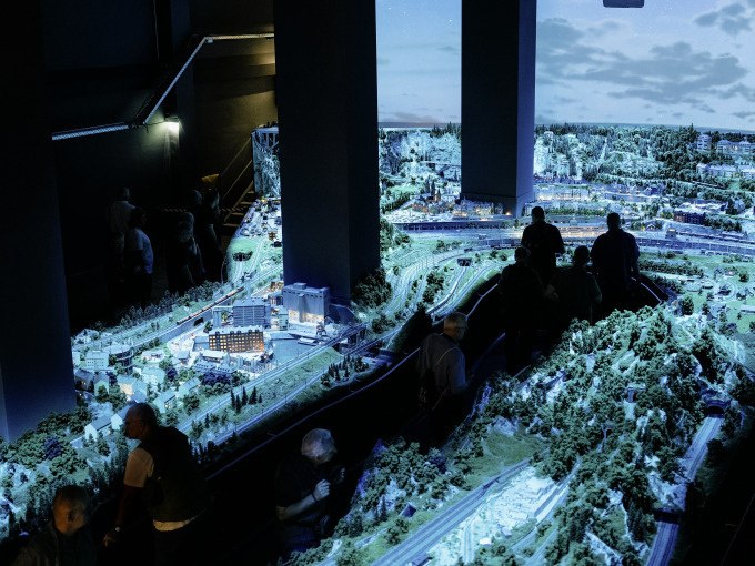 A large model railroad layout in the Märklineum, illuminated in a dark environment, with several visitors looking at the details., © Märklin Spielwaren Vertriebs-GmbH A large model railroad layout in the Märklineum, illuminated in a dark environment, with several visitors looking at the details., © Märklin Spielwaren Vertriebs-GmbH