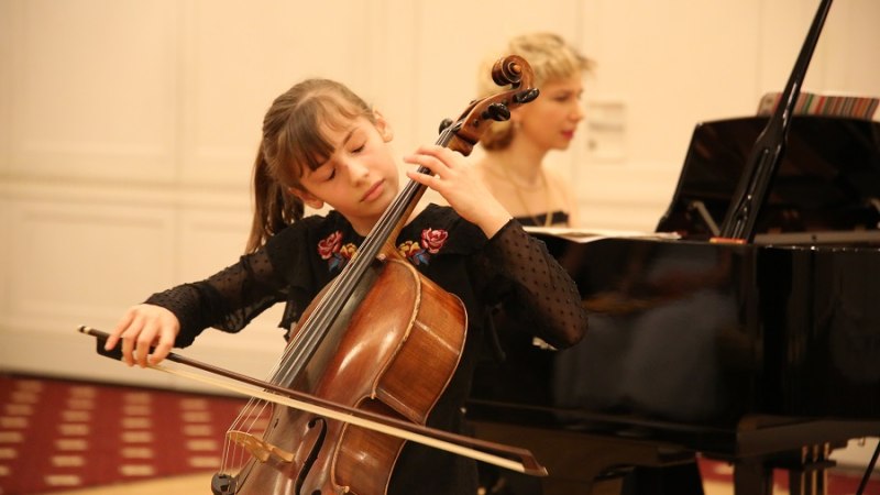 Ein junges Mädchen spielt konzentriert Cello, begleitet von einer Pianistin im Hintergrund in einem eleganten Raum., © Christian Steeneck Ein junges Mädchen spielt konzentriert Cello, begleitet von einer Pianistin im Hintergrund in einem eleganten Raum., © Christian Steeneck