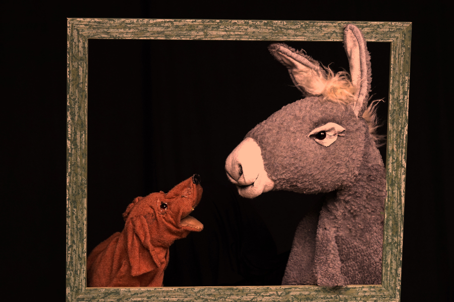 Ein Hund und ein Esel als Handpuppen schauen sich in einem Bilderrahmen an, vor schwarzem Hintergrund., © Theater in der Badewanne Ein Hund und ein Esel als Handpuppen schauen sich in einem Bilderrahmen an, vor schwarzem Hintergrund., © Theater in der Badewanne