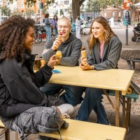 Drei Personen sitzen an einem gelben Tisch im Freien und essen Eis. Im Hintergrund sind Bäume, Fahrräder und ein Café zu sehen., © Stuttgart Marketing GmbH, Sarah Schmid