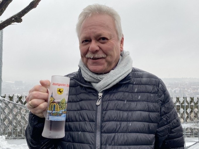 Ein Mann mit grauem Schal und schwarzer Jacke hält einen Glühweinbecher mit Stuttgart-Motiv. Hintergrund: verschneite Landschaft., © Weingenuss Stuttgart Ein Mann mit grauem Schal und schwarzer Jacke hält einen Glühweinbecher mit Stuttgart-Motiv. Hintergrund: verschneite Landschaft., © Weingenuss Stuttgart