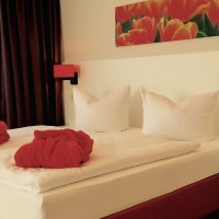 Ein gemütliches Hotelzimmer mit Doppelbett, roten Bademänteln und einem Tulpenbild an der Wand. Die Beleuchtung ist warm und einladend., © TOMAS Ein gemütliches Hotelzimmer mit Doppelbett, roten Bademänteln und einem Tulpenbild an der Wand. Die Beleuchtung ist warm und einladend., © TOMAS