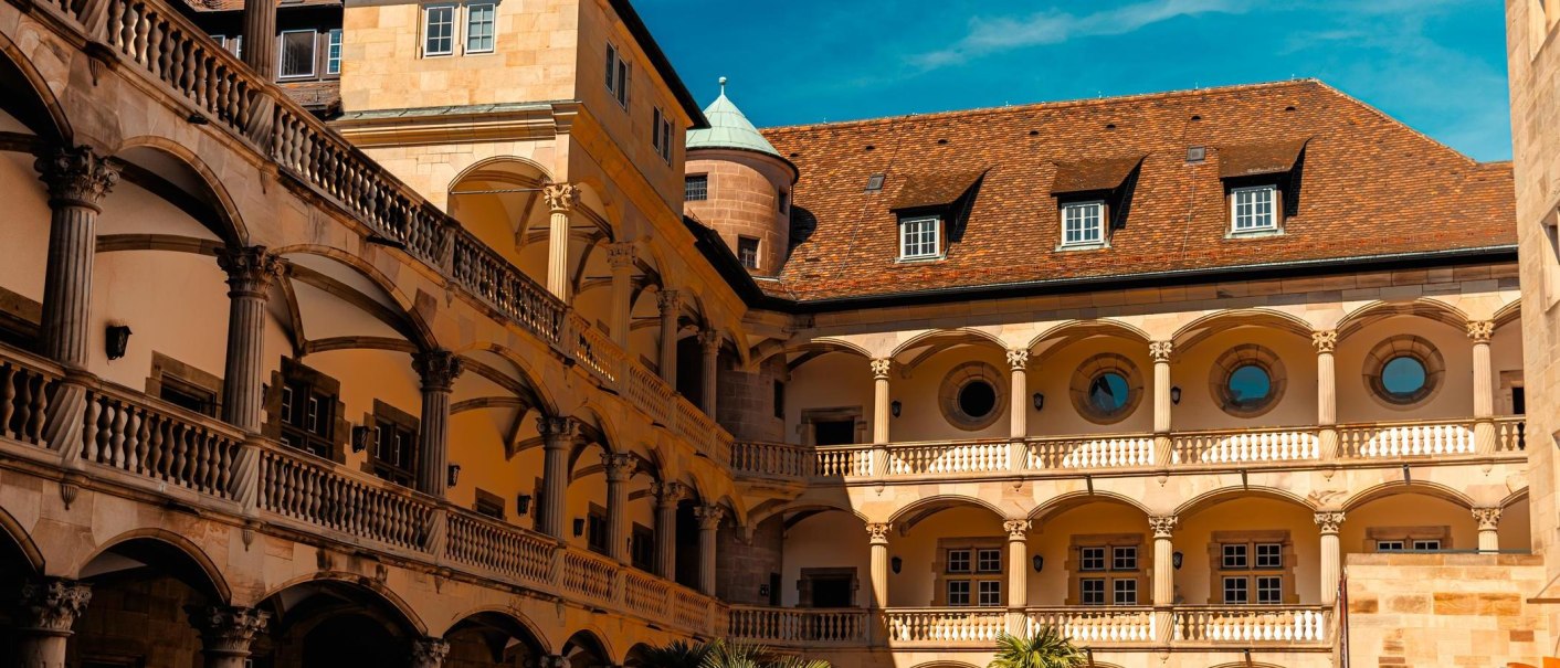 Historischer Innenhof eines Schlosses mit Arkaden, Rundfenstern und einem Turm unter blauem Himmel., © Stuttgart-Marketing GmbH, Sarah Schmid