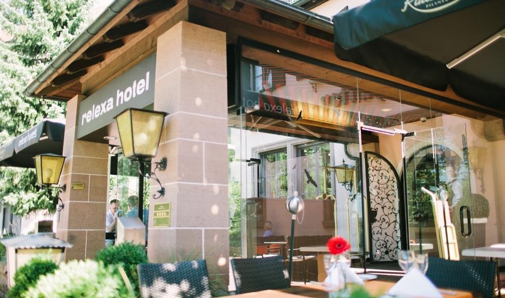 Entrance to the relexa hotel with glass front and terrace. Tables with white tablecloths and flowers, surrounded by green plants., &copy; relexa Waldhotel Schatten