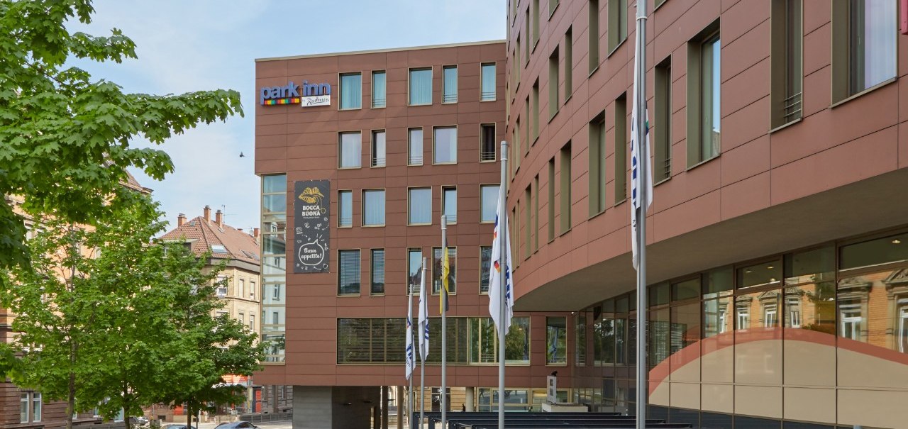 Das Park Inn by Radisson Stuttgart zeigt eine moderne Fassade mit Flaggen davor. Ein Baum und eine Straße sind im Hintergrund sichtbar., © Park Inn by Radisson Stuttgart Das Park Inn by Radisson Stuttgart zeigt eine moderne Fassade mit Flaggen davor. Ein Baum und eine Straße sind im Hintergrund sichtbar., © Park Inn by Radisson Stuttgart