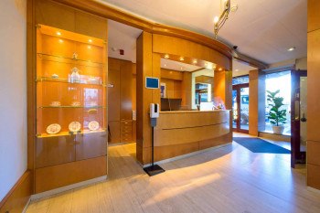 Elegante Hotelrezeption mit Holzverkleidung, Vitrine mit Ausstellungsstücken und einem Empfangstresen. Helle Beleuchtung und Pflanzen im Hintergrund., © PLAZA INN Rieker Stuttgart Hauptbahnhof Elegante Hotelrezeption mit Holzverkleidung, Vitrine mit Ausstellungsstücken und einem Empfangstresen. Helle Beleuchtung und Pflanzen im Hintergrund., © PLAZA INN Rieker Stuttgart Hauptbahnhof