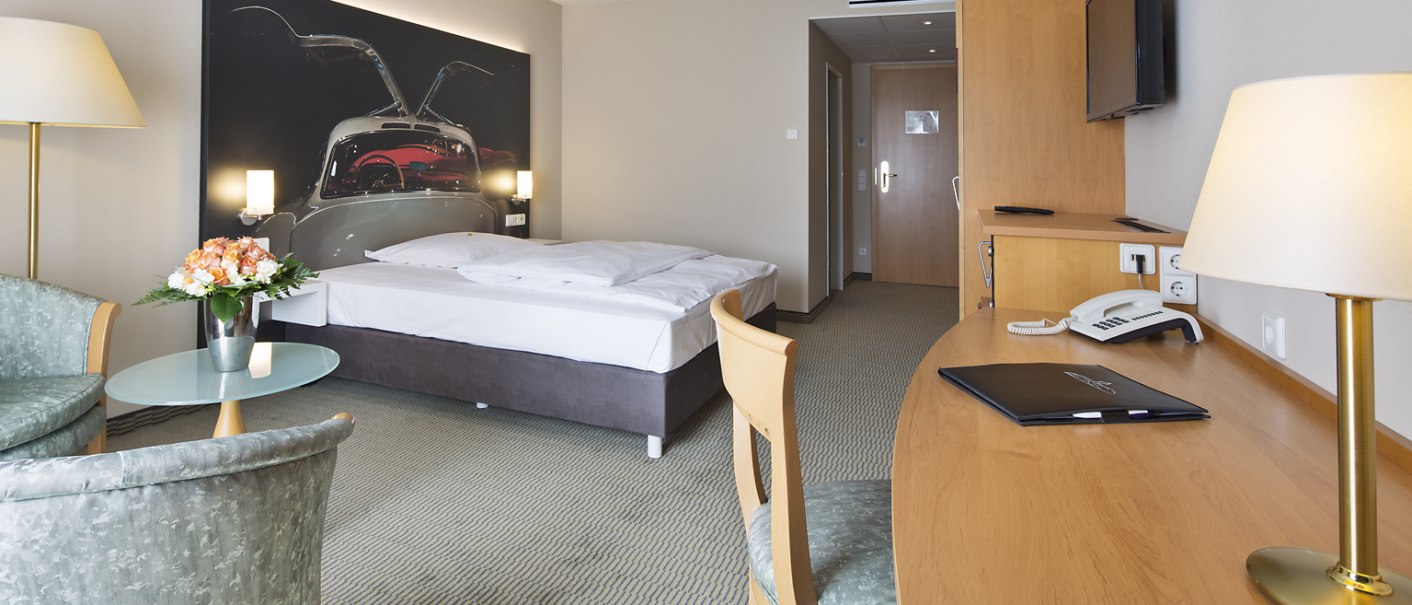 A comfortable hotel room with a double bed, desk, TV and seating area. A large picture of a car adorns the wall behind the bed., © Erikson Hotel