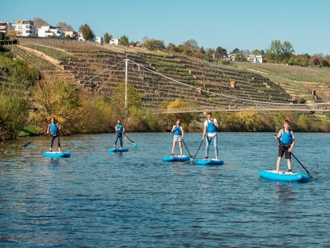 Fünf Personen auf blauen SUP-Boards paddeln auf einem Fluss. Im Hintergrund sind Weinberge und eine Hängebrücke zu sehen., © Cool-Tours StattReisen Fünf Personen auf blauen SUP-Boards paddeln auf einem Fluss. Im Hintergrund sind Weinberge und eine Hängebrücke zu sehen., © Cool-Tours StattReisen