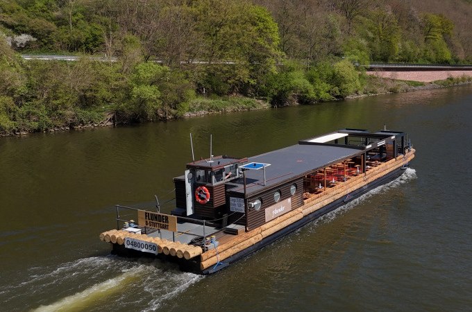 Flussschiff Flunder auf einem Fluss, umgeben von grüner Landschaft. An Bord sind Sitzgelegenheiten für eine BBQ-Tour., © Cool-Tours StattReisen Flussschiff Flunder auf einem Fluss, umgeben von grüner Landschaft. An Bord sind Sitzgelegenheiten für eine BBQ-Tour., © Cool-Tours StattReisen