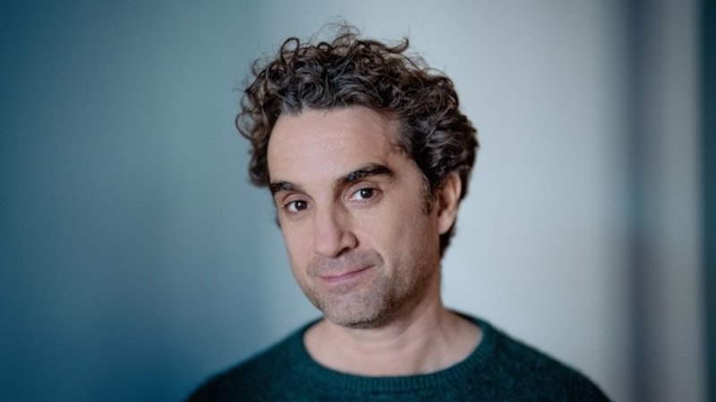 Ein Mann mit lockigem Haar und gr&uuml;nem Pullover blickt leicht l&auml;chelnd in die Kamera. Der Hintergrund ist unscharf und blau-grau., &copy; Renitenztheater Stuttgart e.V.