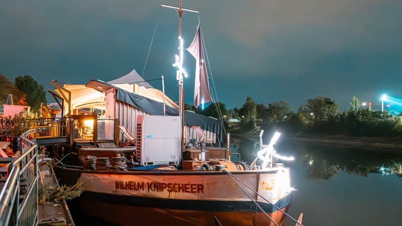 Ein beleuchtetes Boot namens Wilhelm Knipscheer liegt an einem Pier. Im Hintergrund sind B&auml;ume und Lichter zu sehen. Links stehen Tische und St&uuml;hle., &copy; Fridas Pier GmbH
