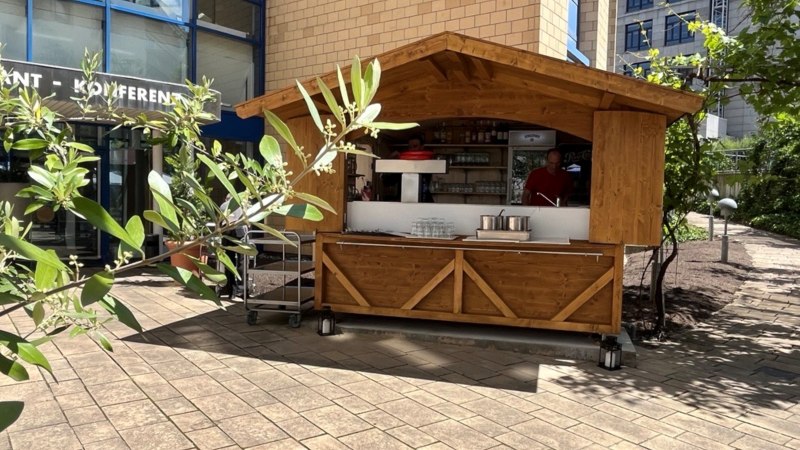 A wooden stand in front of a modern building with a glass fa&ccedil;ade. Plants can be seen in the foreground, partially covering the stand., &copy; TOMAS