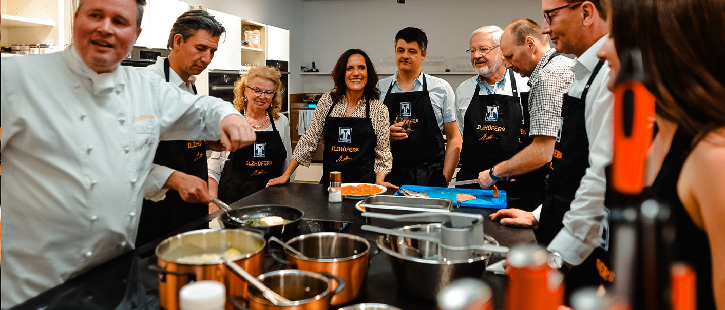 Eine Gruppe von Menschen in einer modernen Küche trägt Schürzen mit 'ILZHÖFER's'. Sie scheinen an einem Kochkurs teilzunehmen., © ILZHÖFER`s Event-Kochschule Eine Gruppe von Menschen in einer modernen Küche trägt Schürzen mit 'ILZHÖFER's'. Sie scheinen an einem Kochkurs teilzunehmen., © ILZHÖFER`s Event-Kochschule