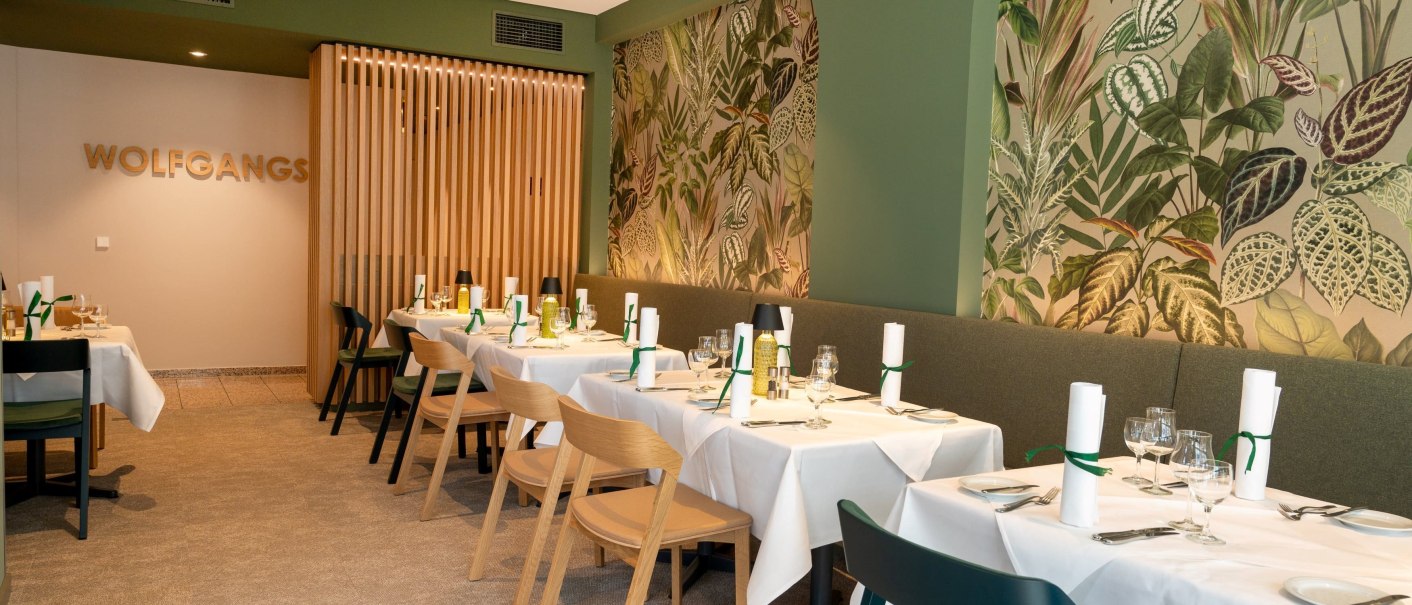Restaurant with green wall and plant motif, tables laid with white tablecloths and wooden chairs, 'WOLFGANGS' lettering on the wall., © Erikson Hotel
