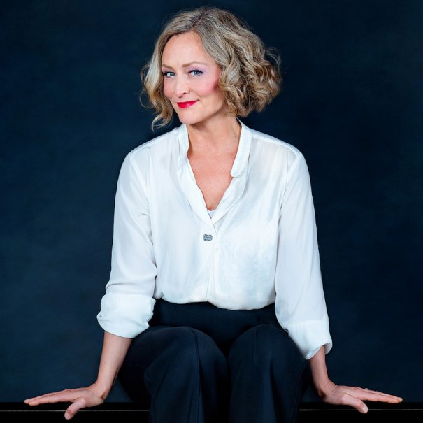 Eine Frau mit blonder Lockenfrisur trägt eine weiße Bluse und schwarze Hose. Sie sitzt lächelnd vor einem dunklen Hintergrund., © Theaterhaus Stuttgart e.V. Eine Frau mit blonder Lockenfrisur trägt eine weiße Bluse und schwarze Hose. Sie sitzt lächelnd vor einem dunklen Hintergrund., © Theaterhaus Stuttgart e.V.