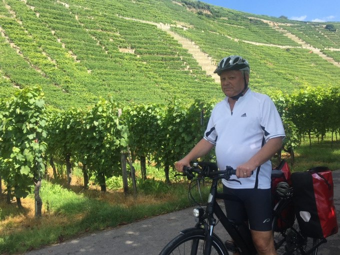 Ein Mann mit Helm steht mit einem E-Bike vor gr&uuml;nen Weinbergen. Er tr&auml;gt ein wei&szlig;es Trikot und hat eine rote Fahrradtasche., &copy; Weingenuss Stuttgart