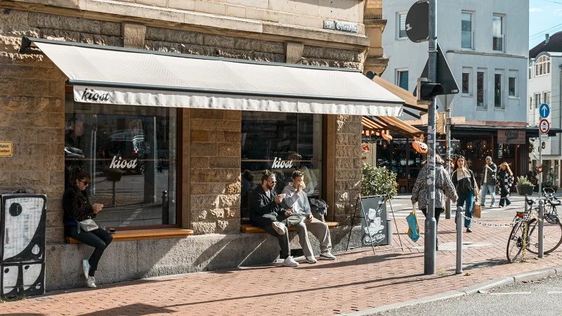 Stra&szlig;enszene vor dem Caf&eacute; Kiost mit Passanten, Fahrr&auml;dern und Sonnenschutz an einer belebten Stra&szlig;enecke., &copy; SMG, Sarah Schmid